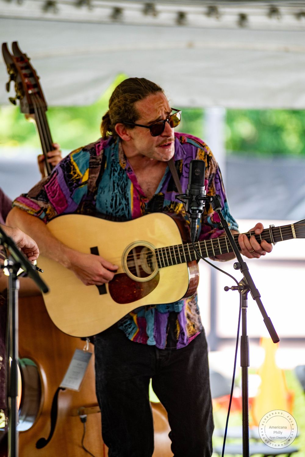 Man in colorful shirt playing acoustic guitar at outdoor festival, wearing sunglasses, performing on stage.
