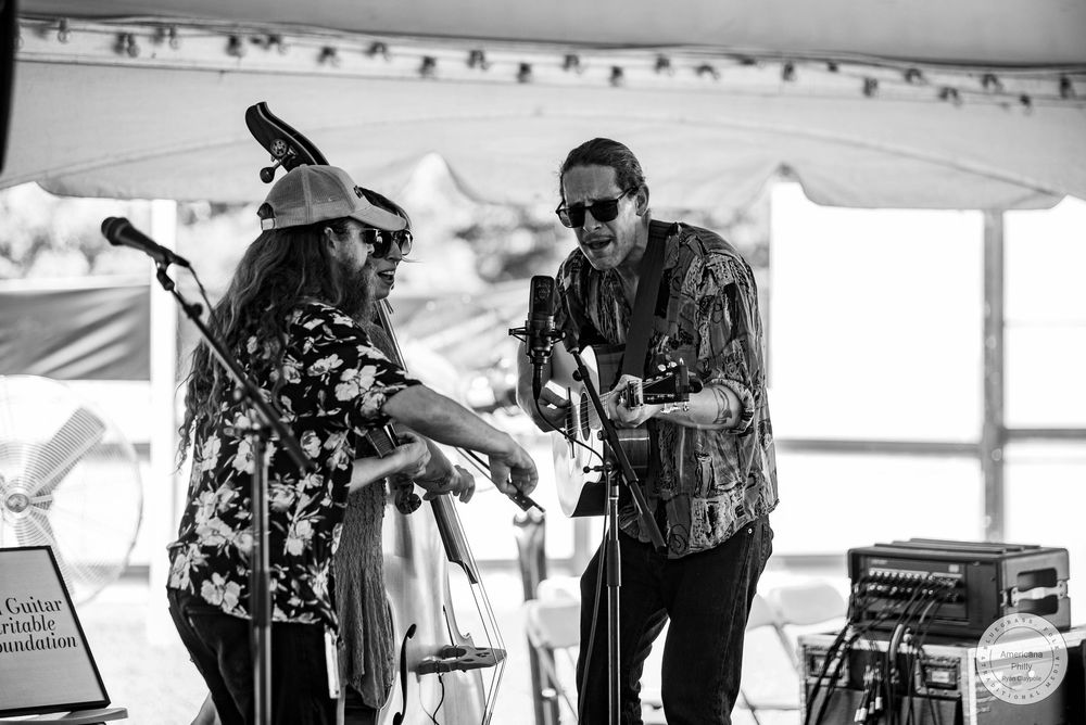 Two musicians perform on stage under a tent, one playing guitar, in a black and white outdoor festival photo.