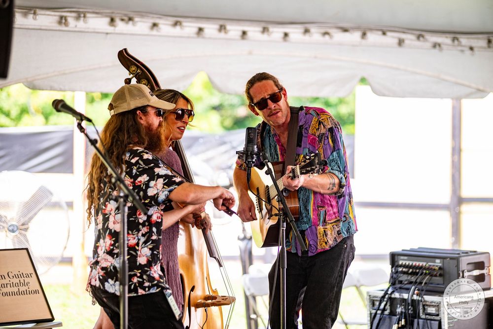Three musicians performing under a tent at an outdoor music festival, with guitars and upright bass.