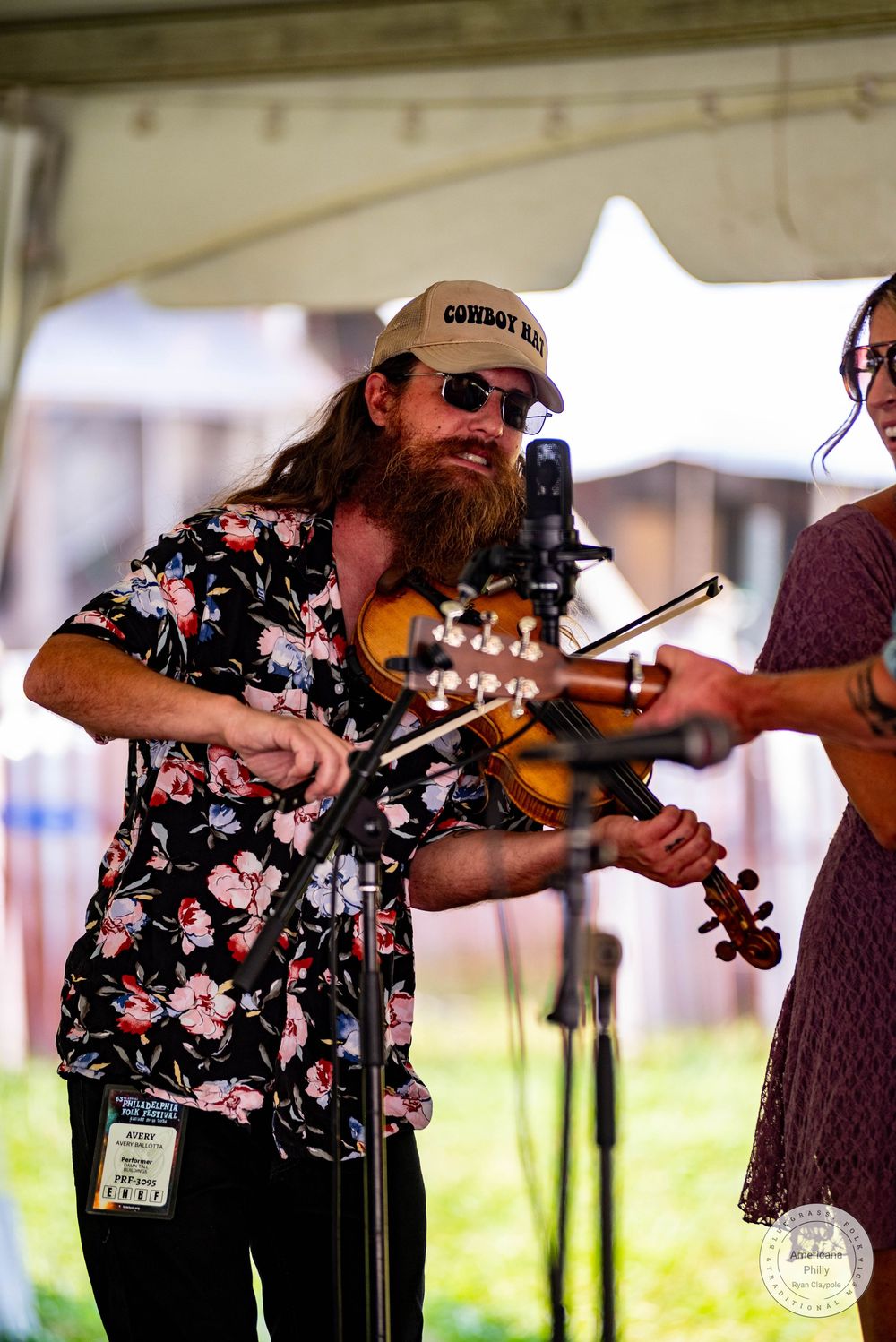 Bearded musician in floral shirt and cowboy hat plays violin on outdoor stage at a music festival.