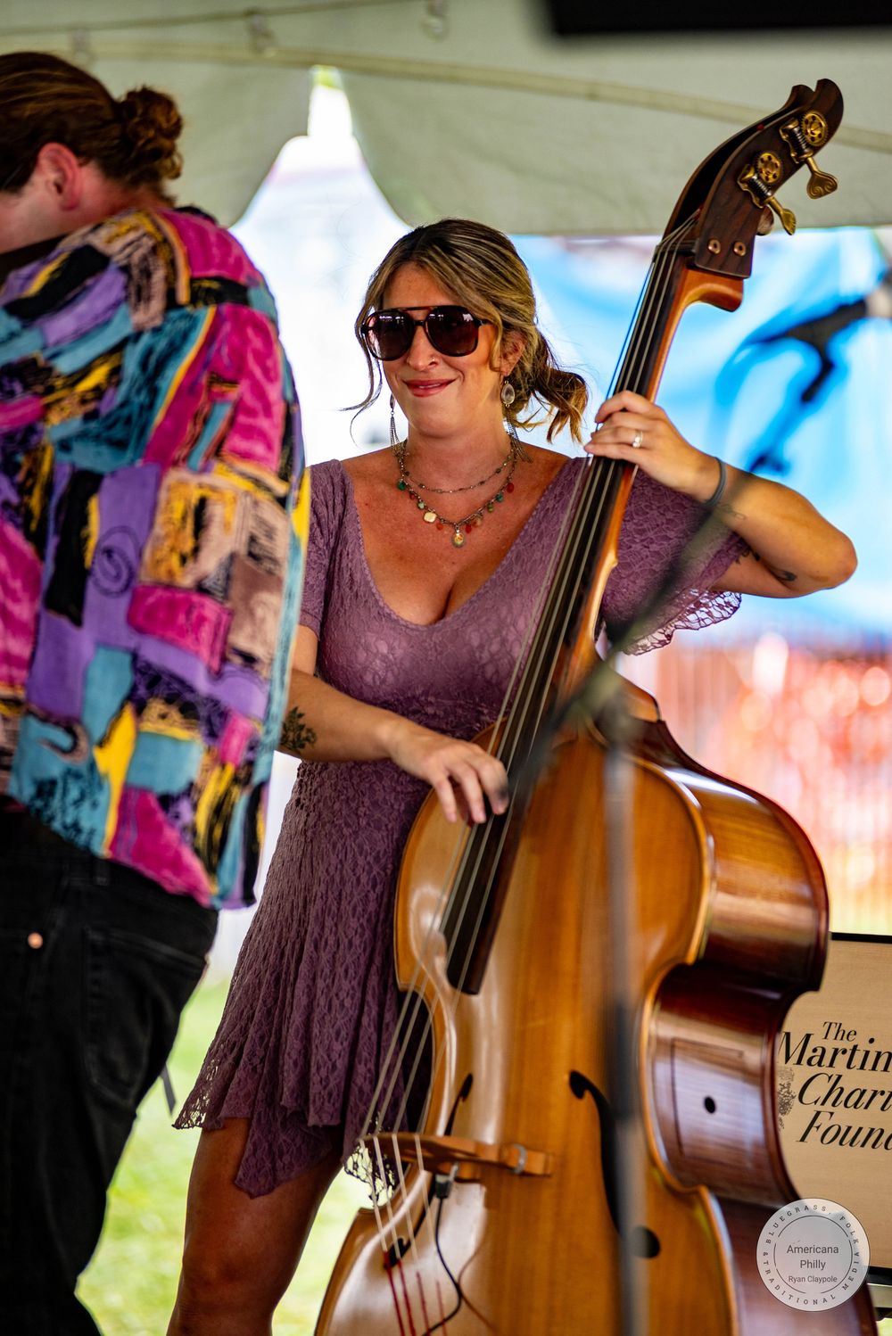 Woman in purple dress playing double bass at outdoor music event, smiling and wearing sunglasses.