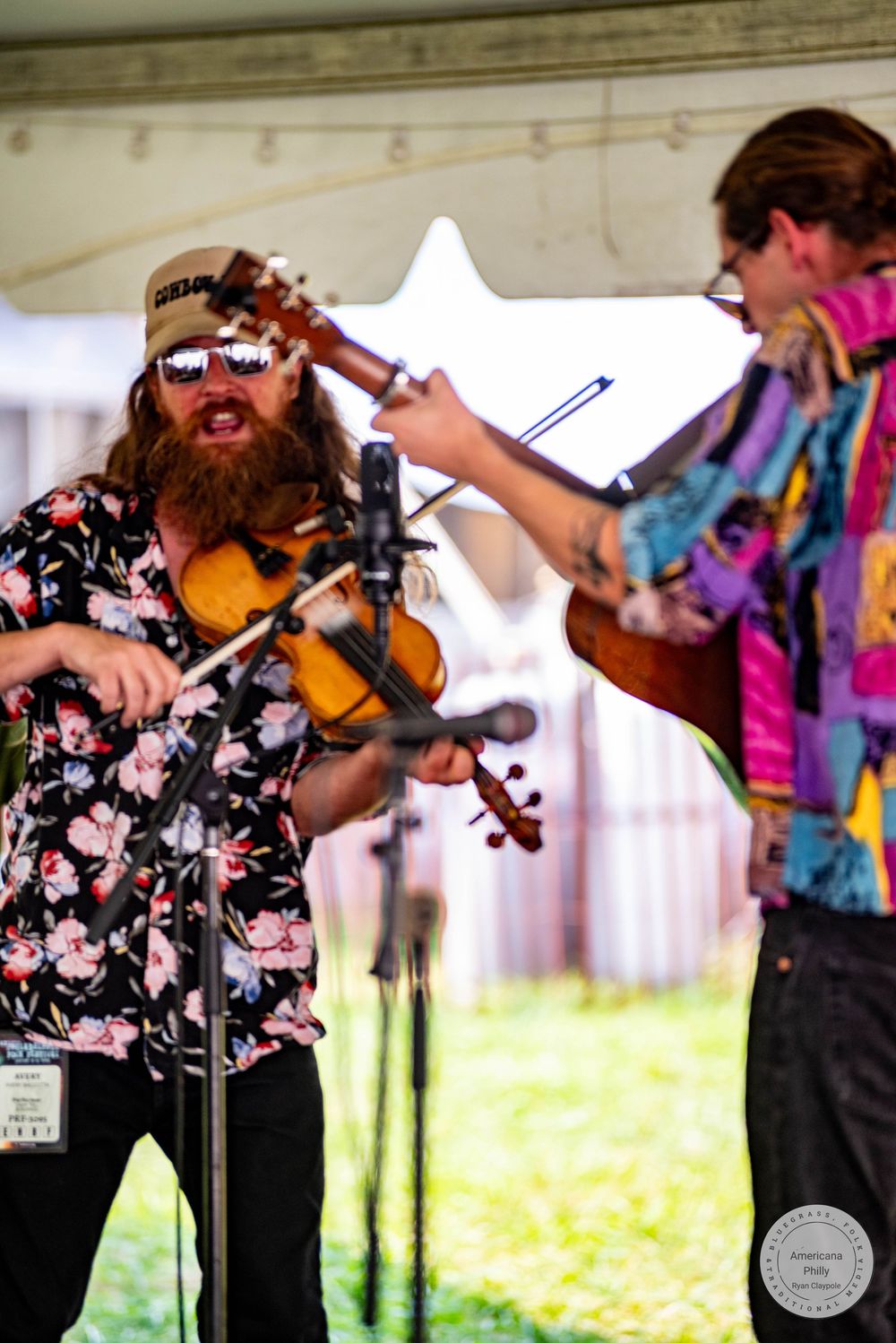 Two bearded musicians play violin and guitar on an outdoor festival stage, wearing colorful shirts under a white tent.