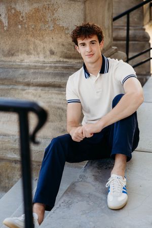 Young man in white polo shirt and navy pants sitting on stone steps wearing white Adidas sneakers.