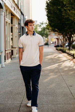Young man in cream polo shirt and navy pants walking on urban sidewalk with trees and storefronts.