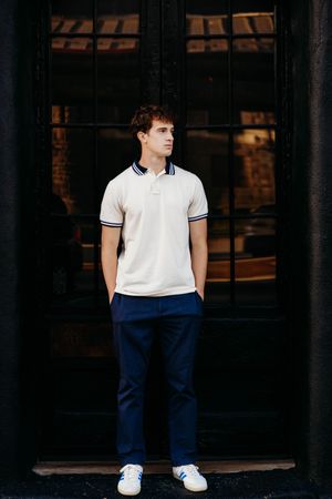 Young man in white polo shirt and navy trousers standing confidently in front of dark glass doors.