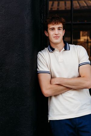 Young man in white polo shirt with arms crossed, leaning against a dark wall, smiling confidently.