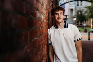 Young man in white polo shirt leaning against a red brick wall outdoors, smiling confidently.