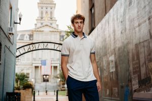Young man in white polo shirt and navy pants standing in urban setting with historic building and arch.