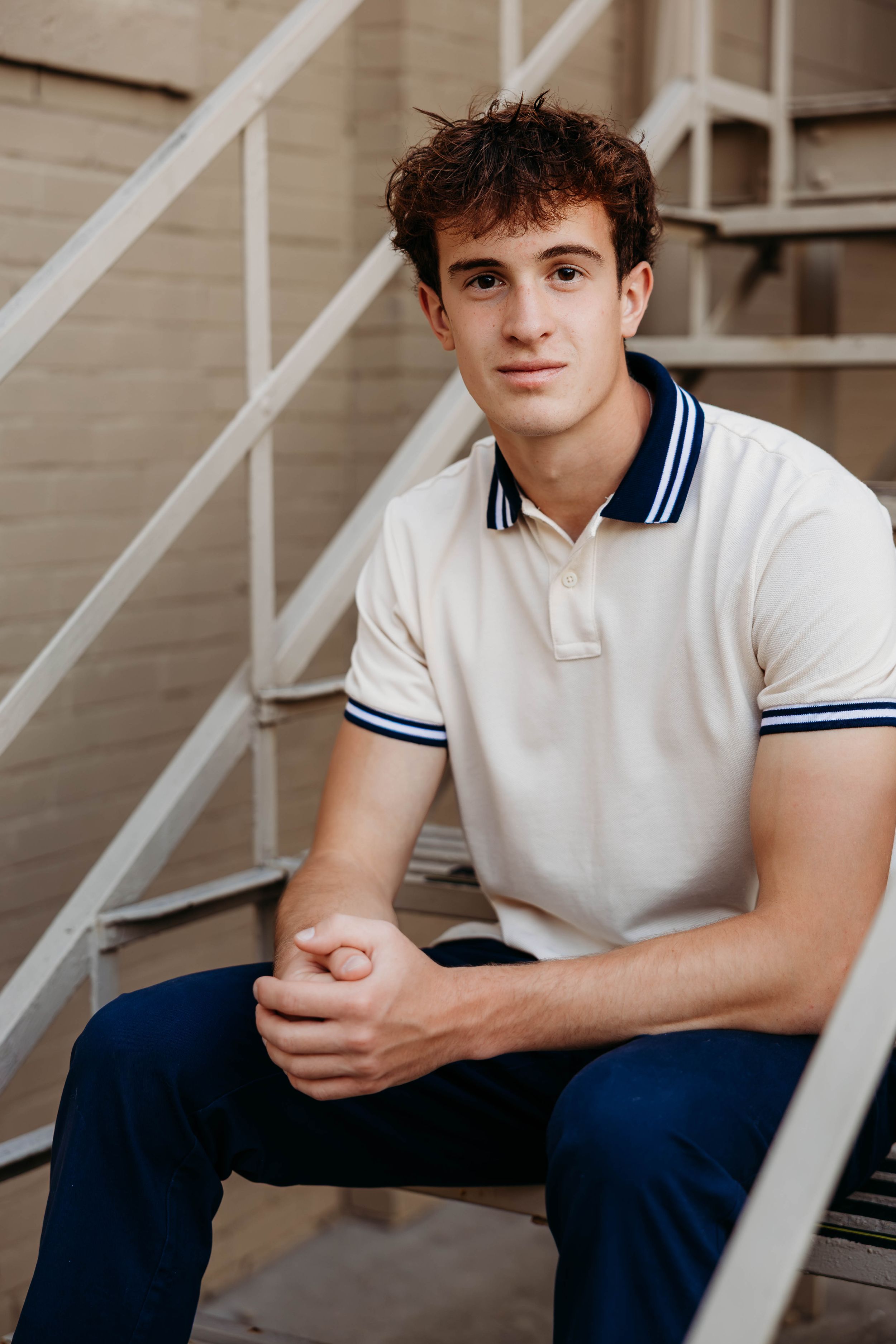 Young man in white polo shirt sitting on metal stairs, smiling confidently at camera.