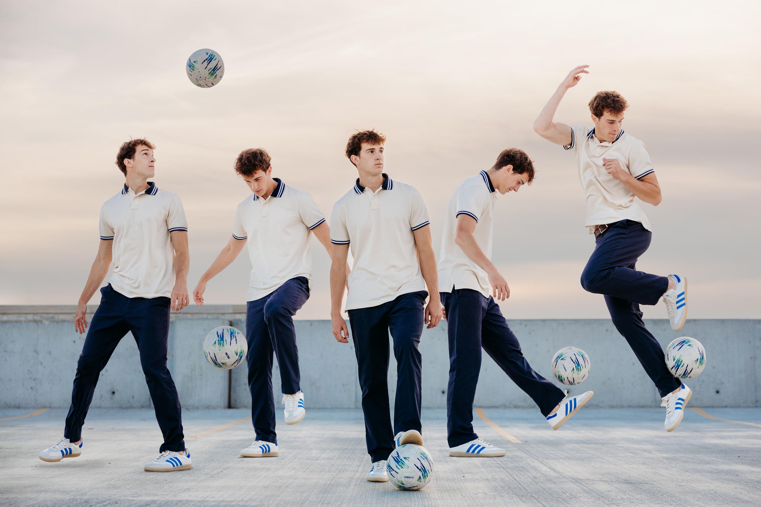 Five young men in white shirts and navy pants play with soccer balls on a rooftop at dusk.