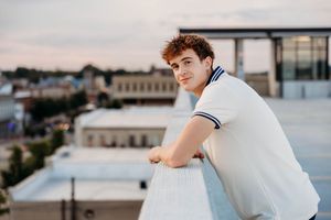 Young man in white polo shirt leaning on rooftop railing at dusk with city skyline in background.
