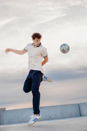 Young man in white polo and navy Adidas pants juggling a soccer ball outdoors under a cloudy sky.