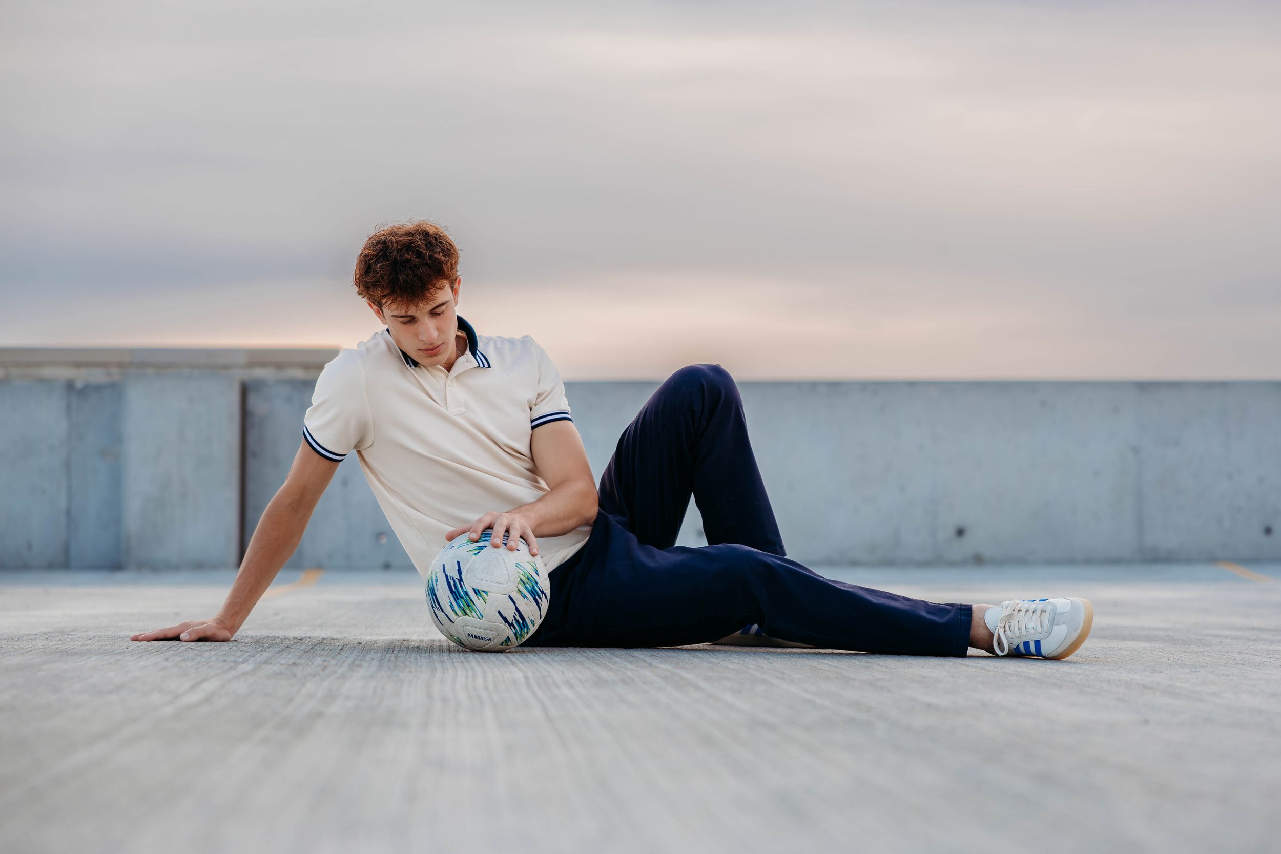Young man sitting on rooftop floor holding a soccer ball, wearing white polo shirt and navy jeans.