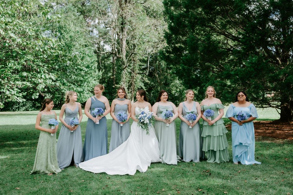 Bride in white gown stands with bridesmaids in sage green dresses holding blue bouquets outdoors.