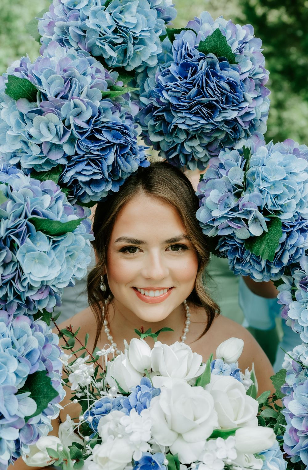Smiling bride surrounded by blue and white hydrangea flowers holding a bouquet on her wedding day.