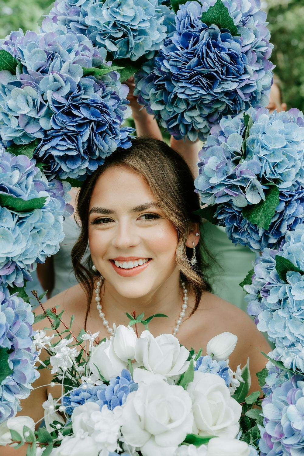 Smiling bride surrounded by blue hydrangea bouquets holding white roses, wearing pearl necklace and earrings on wedding day.