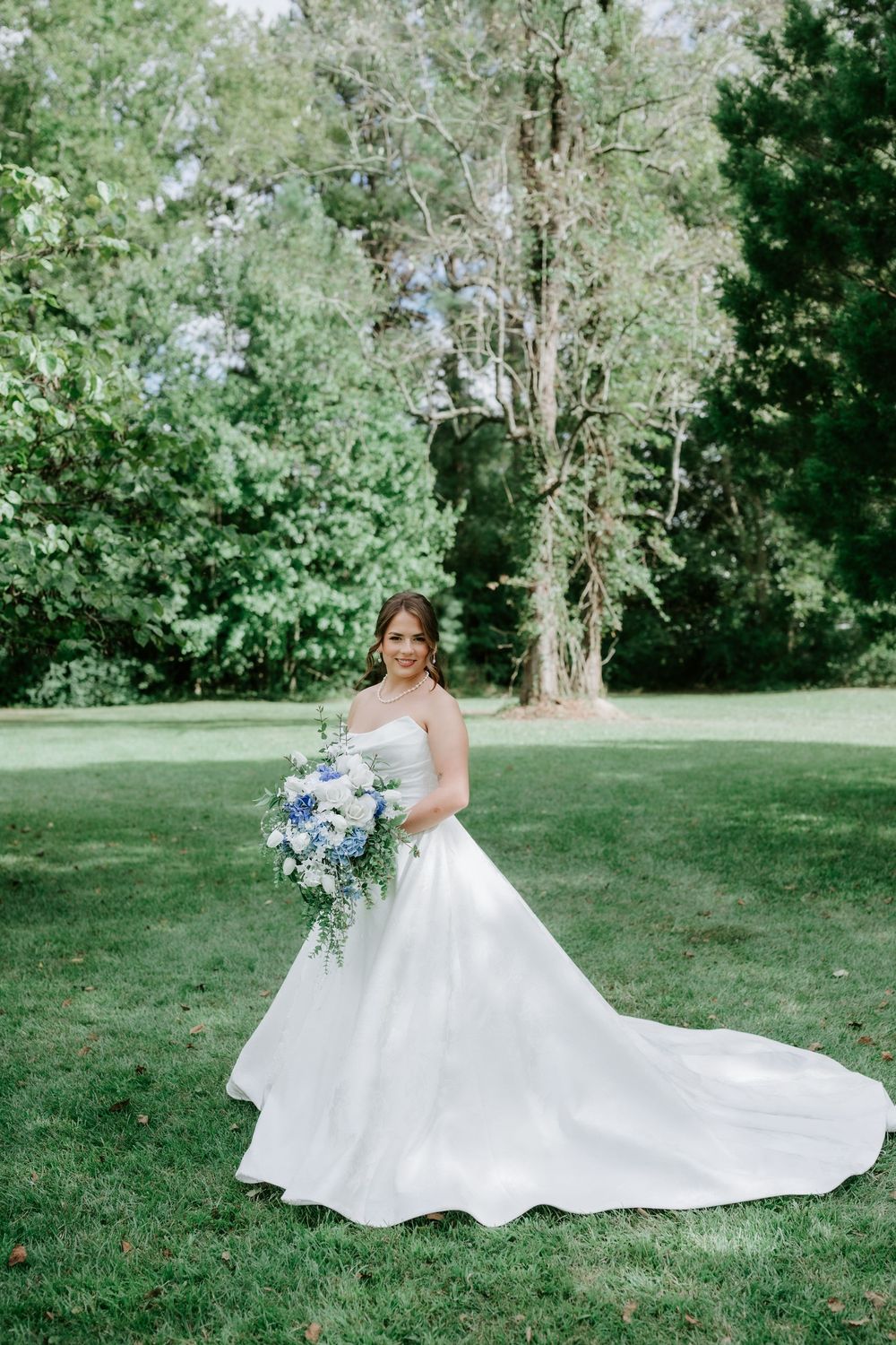Bride in a strapless white ball gown holding a blue and white floral bouquet, standing on green grass with trees behind.