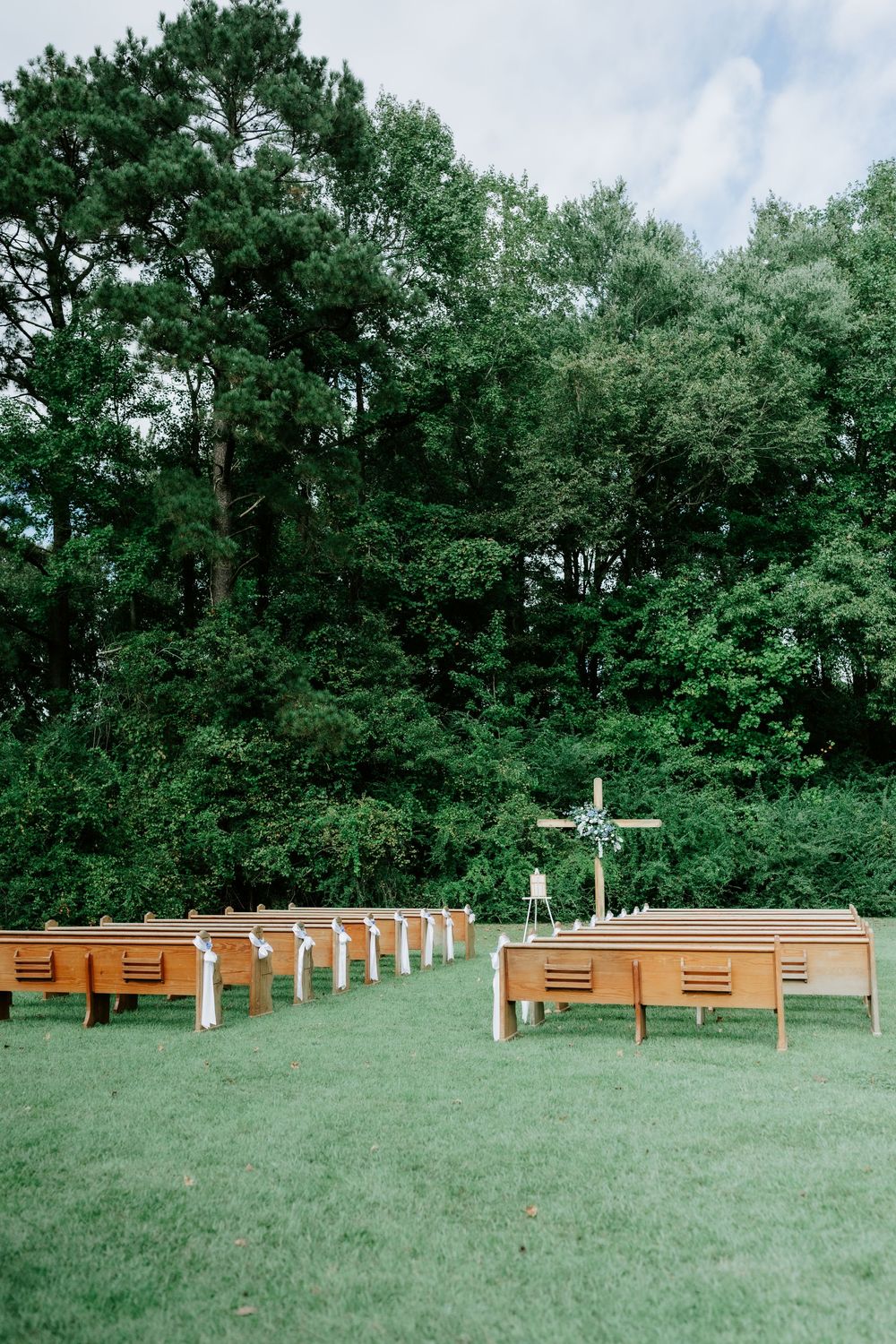 Outdoor wedding ceremony setup with wooden pews on green grass surrounded by tall trees.
