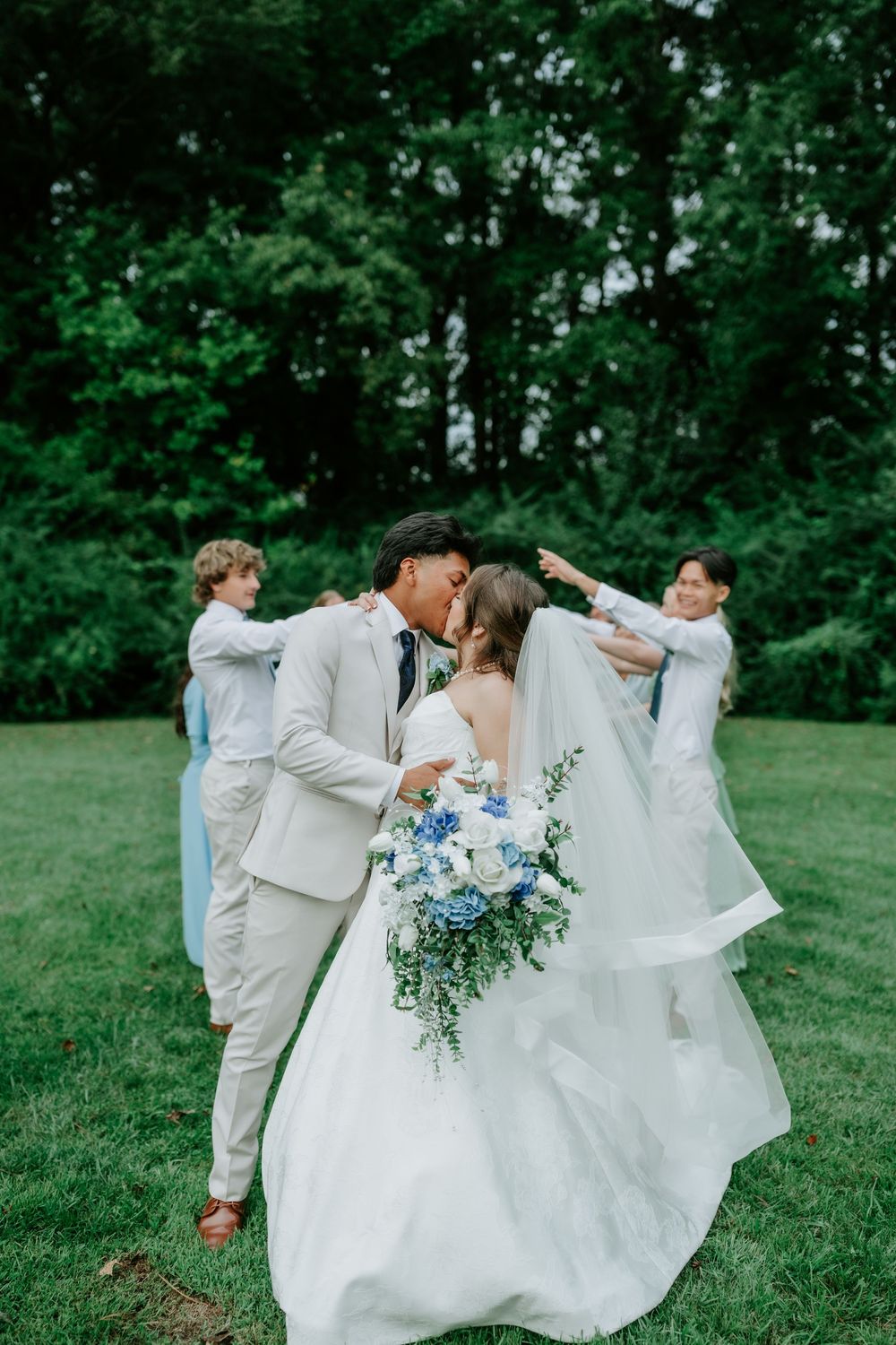 Bride and groom share a kiss outdoors while two boys hold up the wedding veil on a lush green lawn.