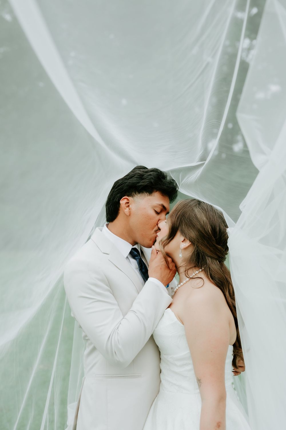Bride and groom share a romantic kiss under a flowing white veil on their wedding day.