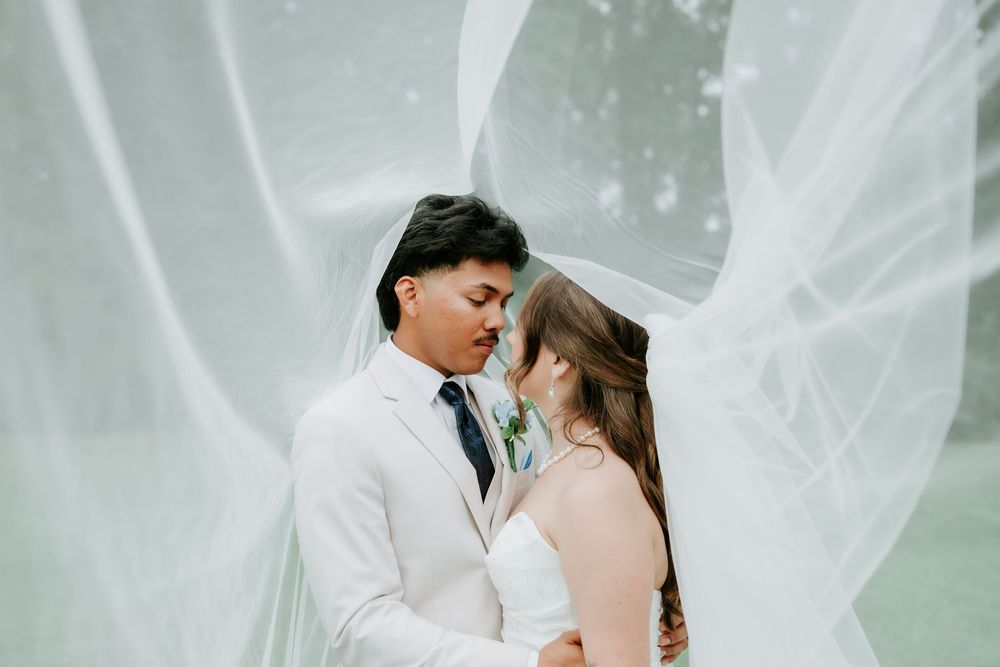 Bride and groom share a romantic moment under a flowing white veil outdoors on their wedding day.