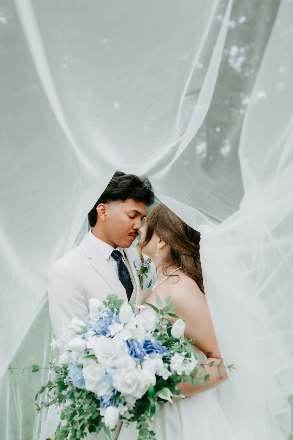 Bride and groom share a romantic moment under flowing white veil, bride holding blue and white floral bouquet.