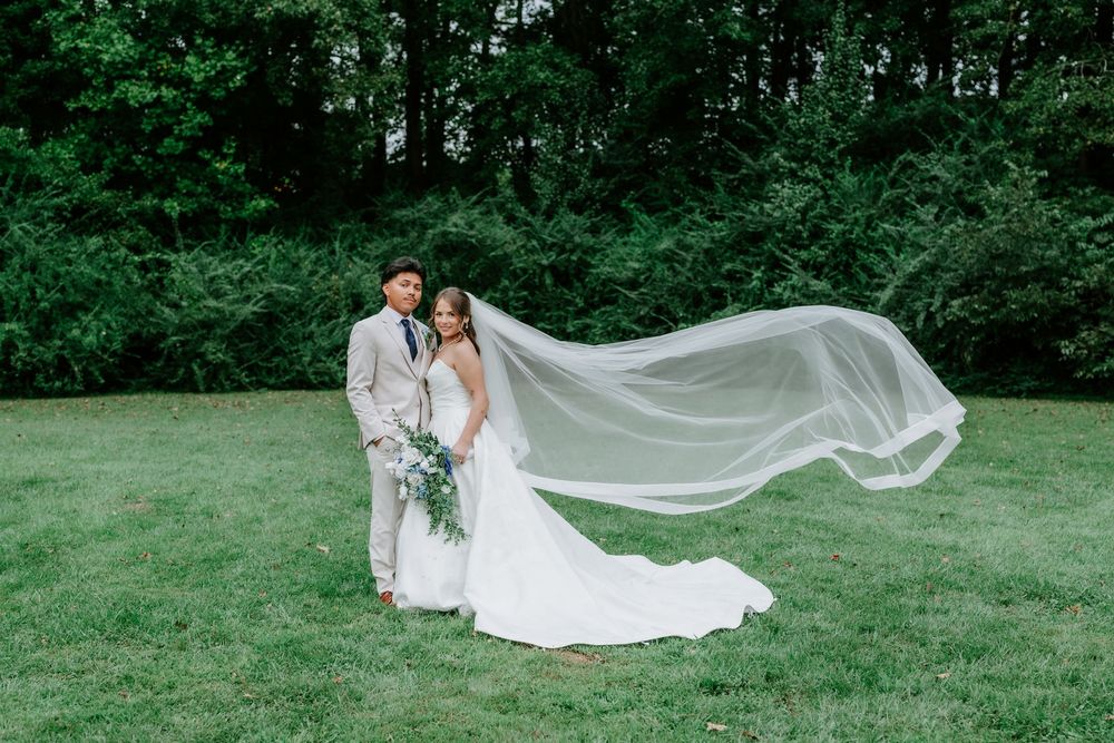 Bride and groom pose on green lawn, her veil flowing in wind, surrounded by lush trees on their wedding day.
