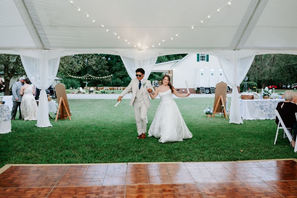Bride and groom make grand entrance at outdoor wedding reception under white tent with string lights.
