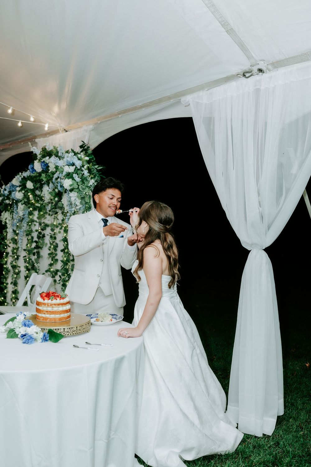 Bride and groom cut their floral-adorned naked wedding cake under a white tent at night.