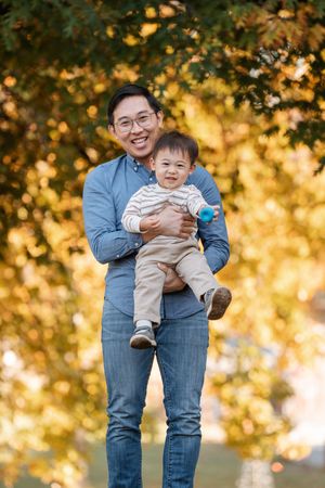 Smiling Asian man holding happy baby boy outdoors among golden autumn trees.