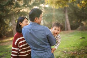 Asian family with parents and baby enjoying outdoor park time, father holding laughing toddler in sunny green setting.