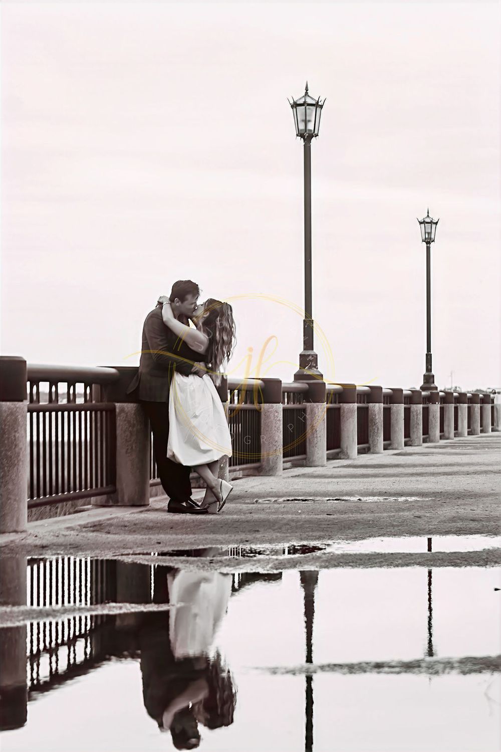 Couple embracing on a waterfront promenade with street lamps, reflected in puddles below in a romantic black and white scene.