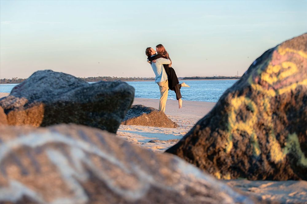 A couple embraces on a rocky beach at sunset with calm blue water in the background.