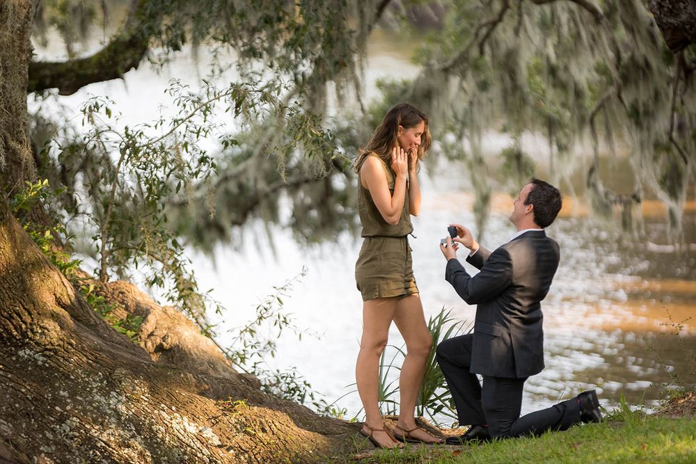 Man proposes to woman on one knee by a lake under a tree, presenting a ring box in an outdoor setting.