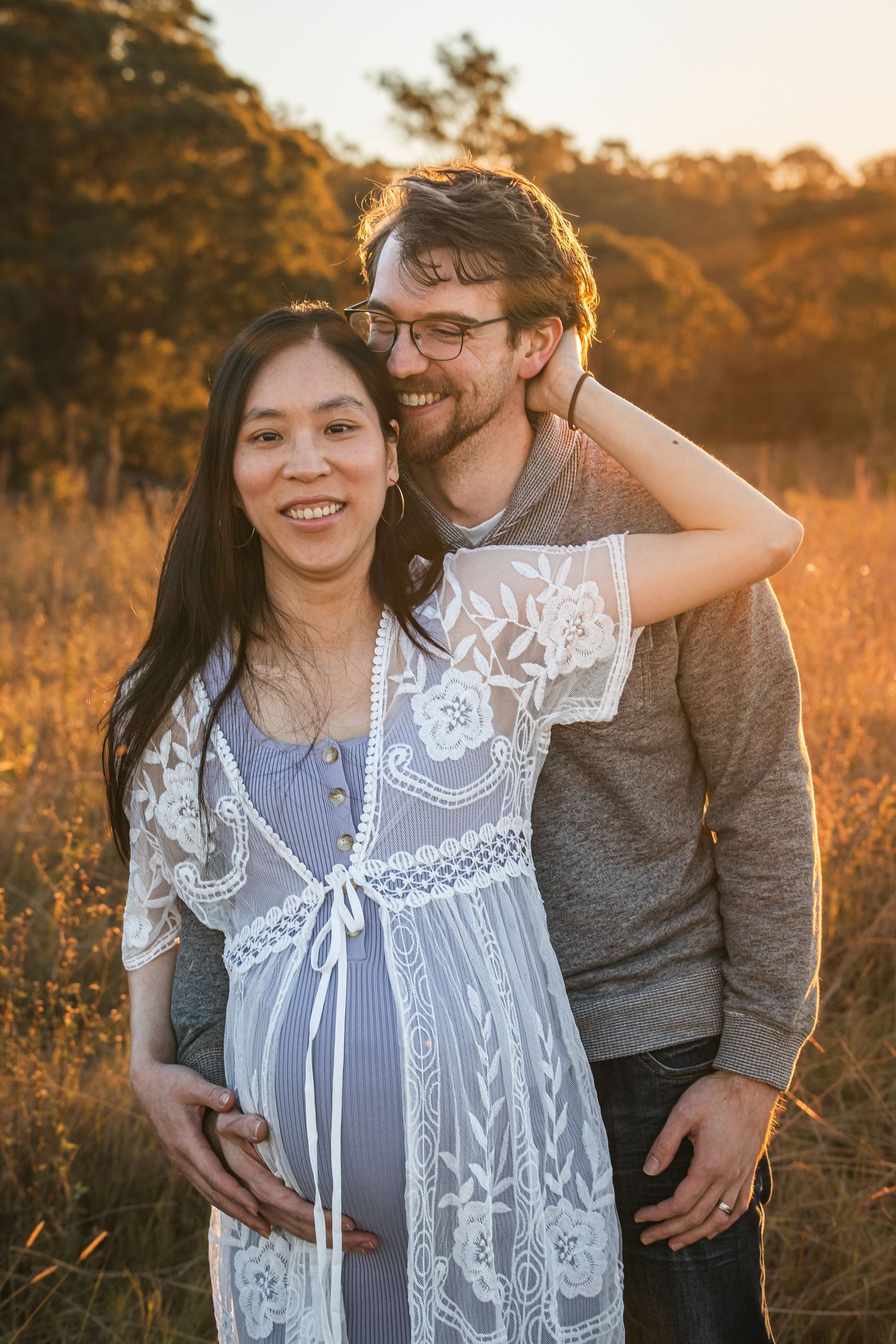 Smiling couple poses in golden field at sunset in Rouse Hill, pregnant woman in white lace dress with partner embracing her bump.