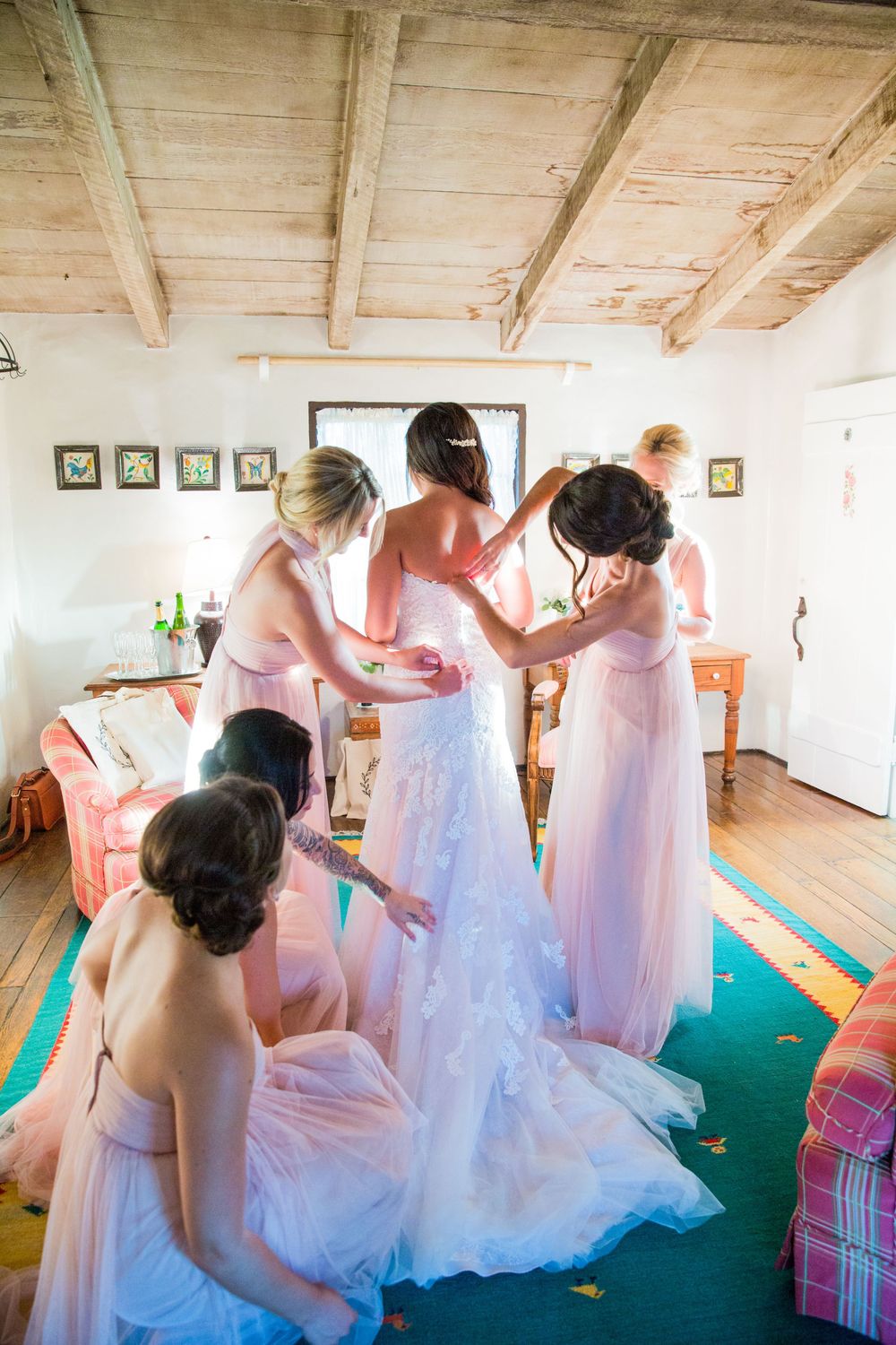 Bridesmaids in blush pink gowns help bride into her white lace wedding dress in a rustic bridal suite.