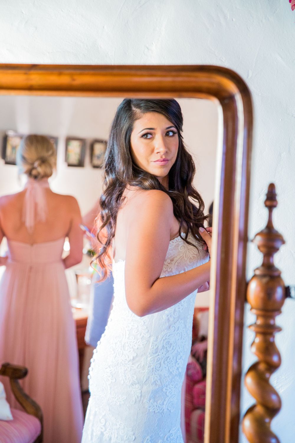 Beautiful dark-haired bride in strapless lace gown gazes over shoulder in ornate gold-framed mirror.