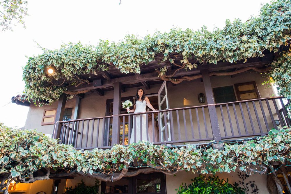 Bride in white gown standing on ivy-covered balcony holding bouquet at rustic wedding venue.
