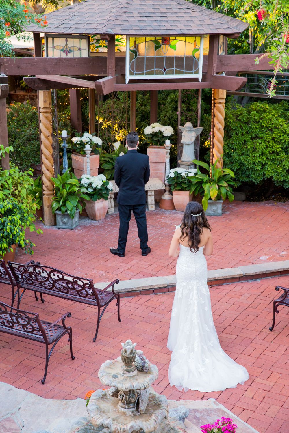 Bride and groom share first look moment in brick courtyard near wooden gazebo with white flowers.