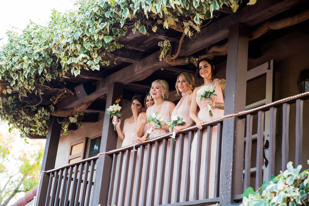 Four bridesmaids in blush dresses holding bouquets pose on a vine-covered wooden balcony outdoors.