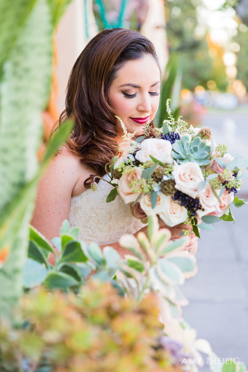 Beautiful bride gazes down at her bouquet of roses, succulents, and wildflowers in soft natural light.