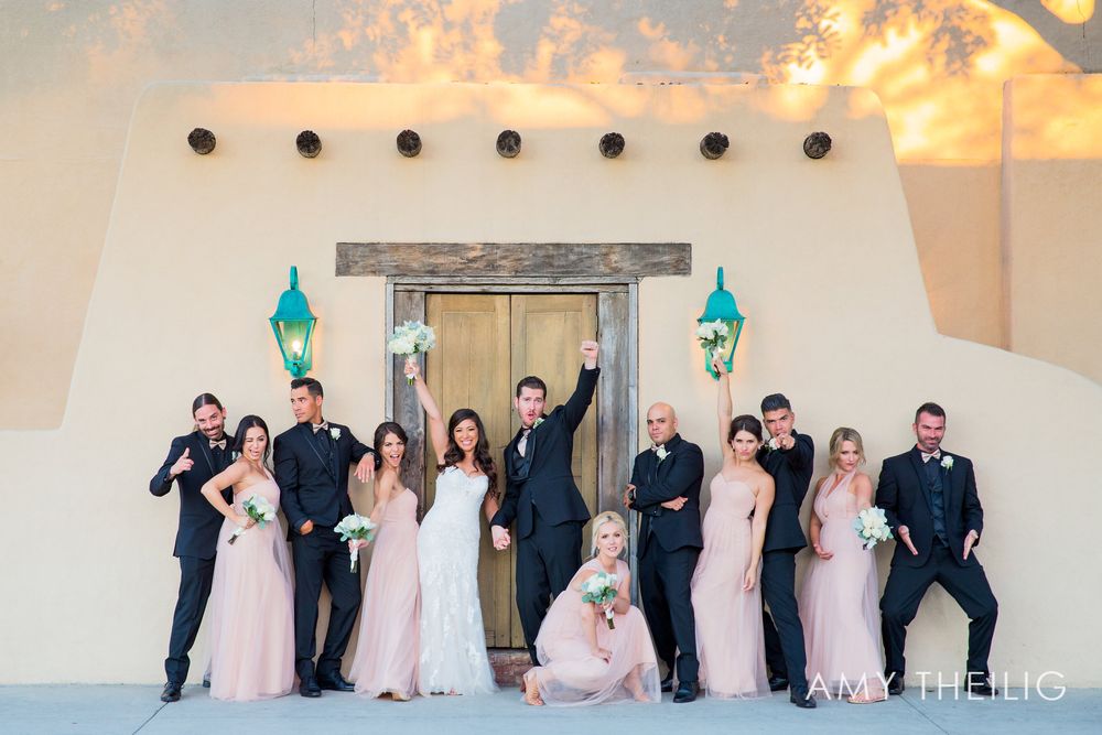 Wedding party in pink bridesmaid dresses and black suits celebrating outside rustic adobe venue.