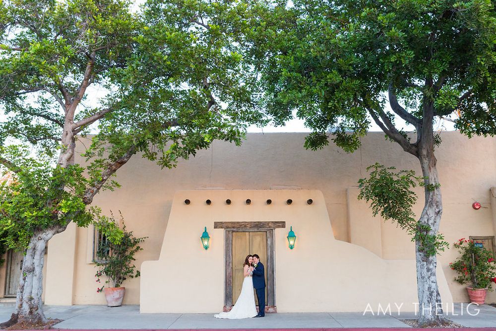 Bride and groom share a kiss outside a white adobe wedding venue framed by large shade trees.