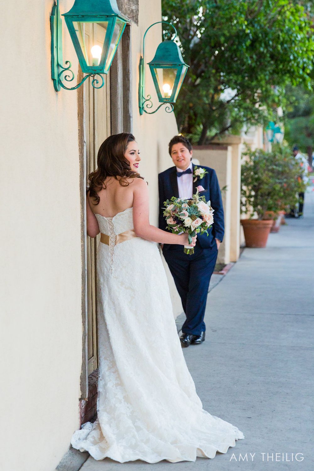 Bride in strapless lace gown smiling as groom approaches along elegant outdoor corridor.