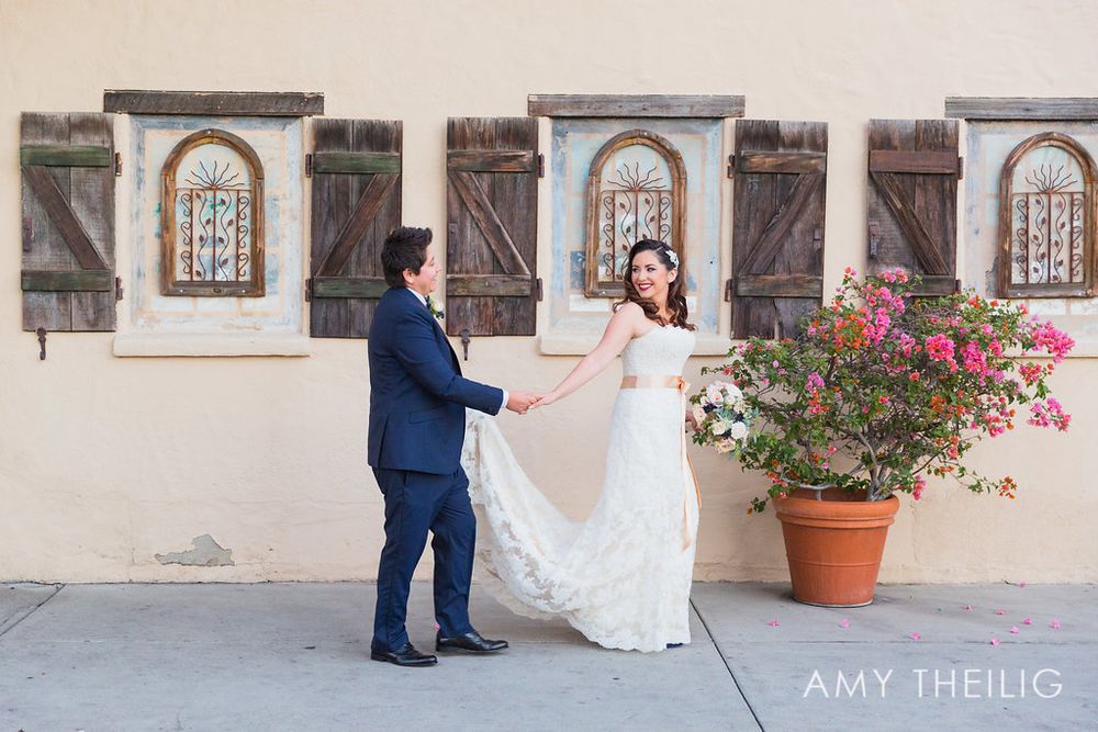 Bride and groom hold hands near a wall with ornate wooden shuttered windows and pink bougainvillea.
