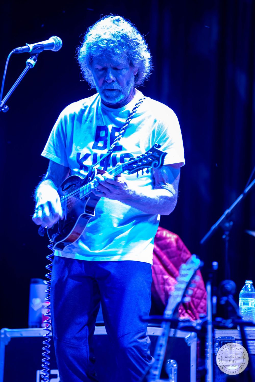 Guitarist in a white Be Kind t-shirt performs on stage under blue lighting at a live concert.