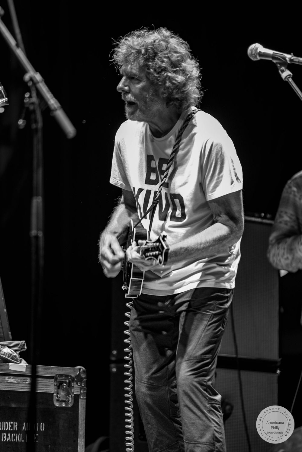 Black and white photo of a curly-haired guitarist in a Be Kind shirt performing passionately on stage.