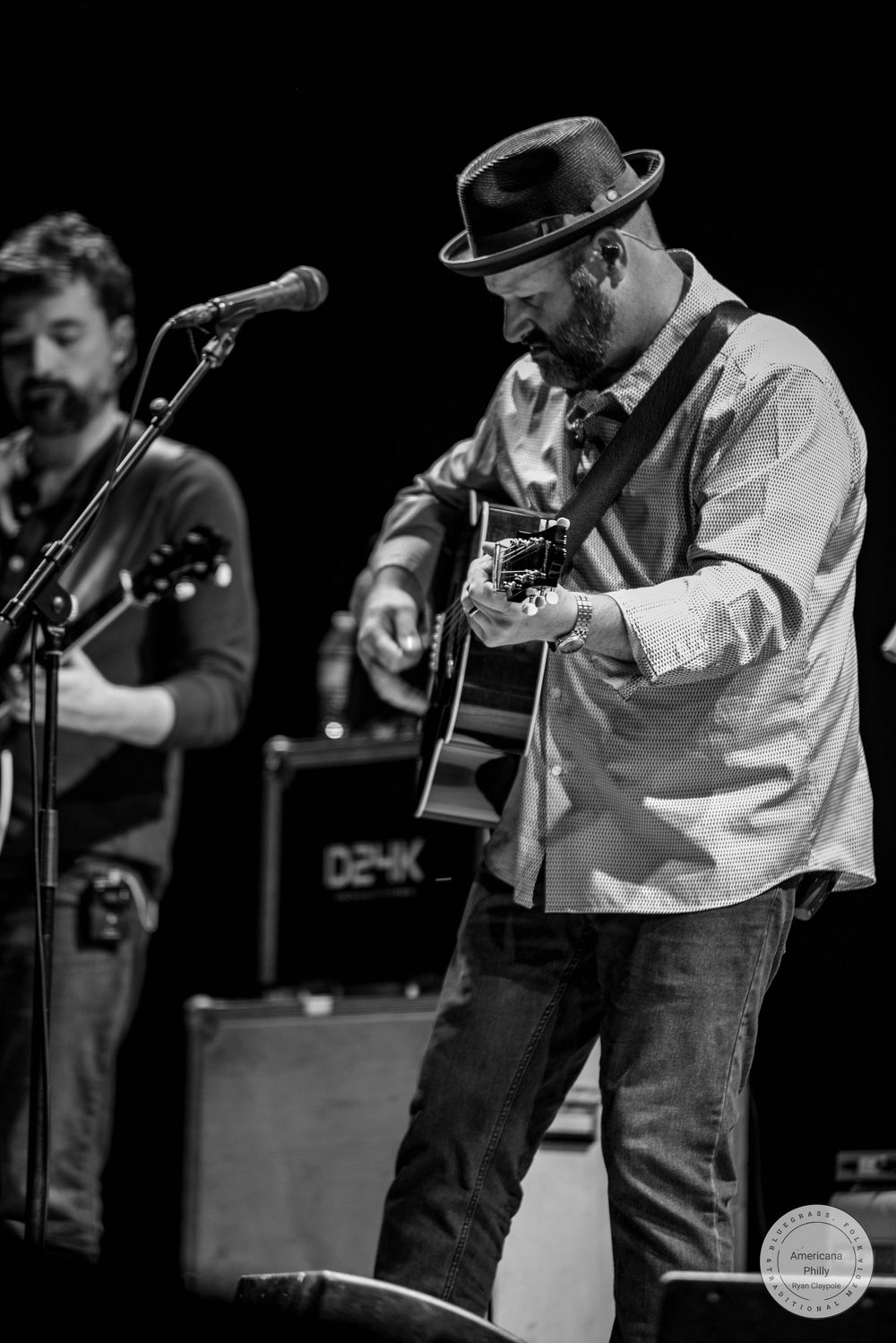 Black and white photo of two male musicians performing on stage, one playing acoustic guitar wearing a hat and casual shirt.