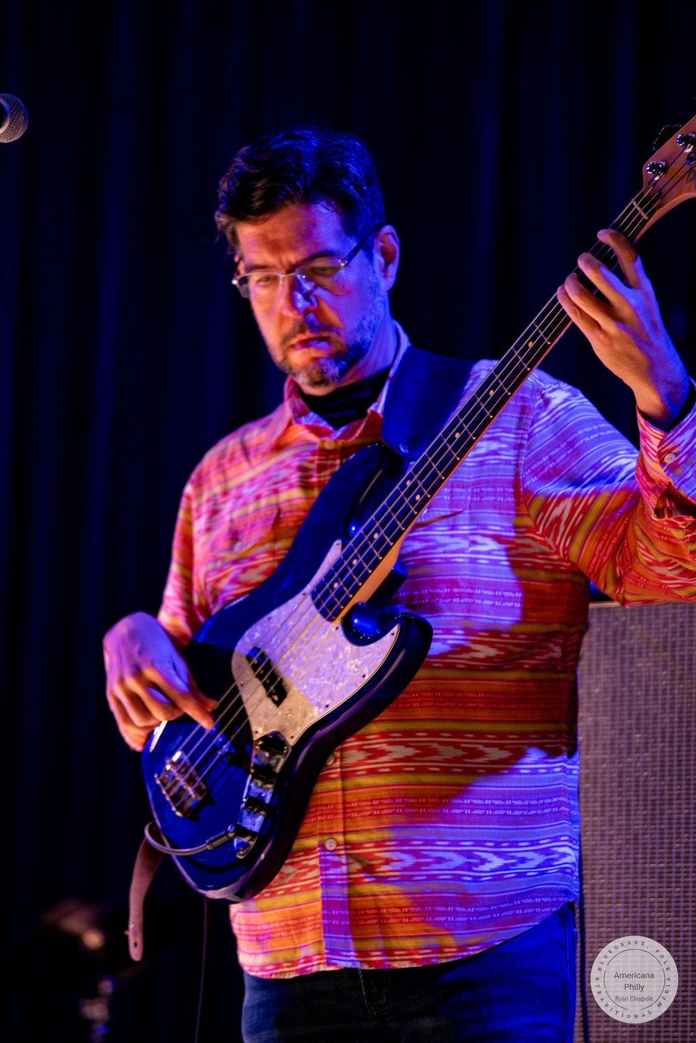 Musician playing a black bass guitar on stage, wearing a colorful patterned shirt under blue and orange stage lighting.