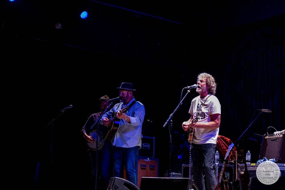 Two musicians perform on stage, one playing guitar and another at a microphone during a live concert.